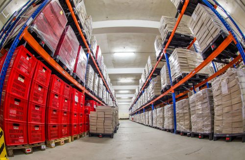 Floor-to-ceiling shelves of red crates and cream-colored boxes wrapped in clear plastic are shown in a well-lit storage room.
