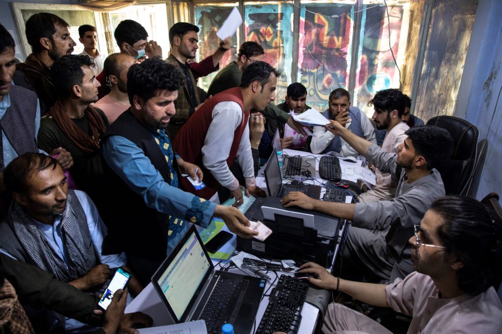 A panicked crowd presses against a desk with several people sitting behind computers in a small room.