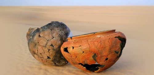 One brown and one orange pot with cracks and missing fragments lean against each other in the sand.