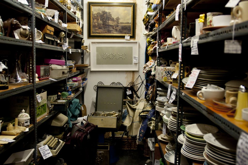 Two shelving units with multiple levels are packed with an assortment of objects. More objects are on the ground below two paintings hung on the back wall.