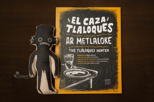 A black stuffed figure with large eyes and a wide mouth wearing a black hat lies beside a black, white, and yellow comic book on a dark wooden table.