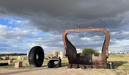 A large black tire and rusted metal mining equipment sit on brown grass under a cloudy sky.