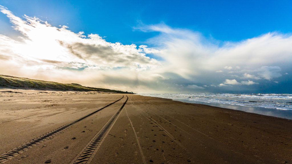 The image shows a wide shot of tire tracks going straight down the sand on a beach underneath a bright blue sky.