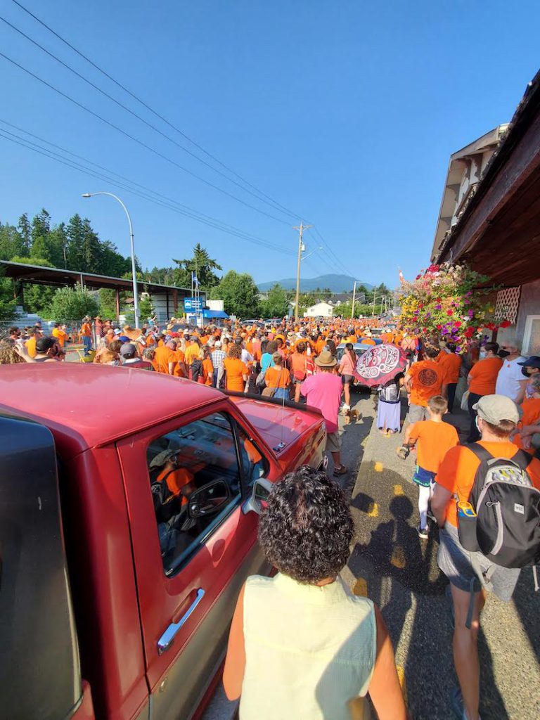 A crowd of people wearing orange shirts walks down a wide street. A red truck rolls alongside those marching.