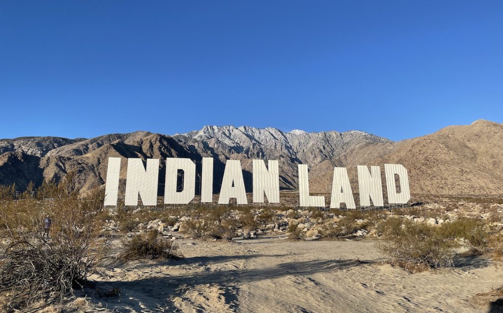 Large white letters that spell out “Indian Land” rest on sand and dried grasses in front of a mountain range.