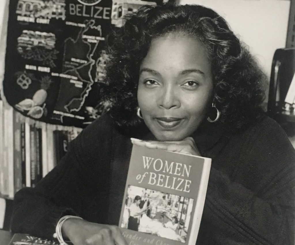 A black-and-white photograph of a woman at a desk in front of a shelf of books features her resting her chin on an upright book, with other books lying flat on the table.