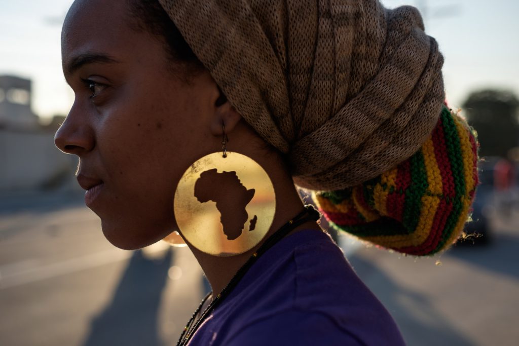 This profile shows a young protestor wearing a large, round, gold earring with a cutout of the African continent and two multicolored headwraps.