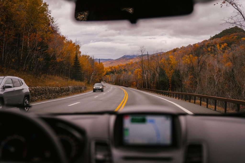 The view is of rolling hills covered with yellow- and red-leafed trees seen through the front window of a car on the highway.