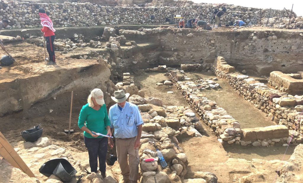 Two people wearing hats look at papers while standing among stones, dirt, and short, ancient walls.