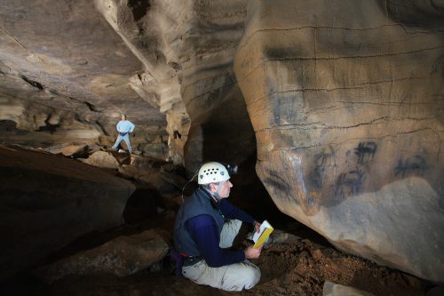 A person wearing a white helmet sits on the floor of a cave holding a yellow notebook and looking at drawings on a wall. Another person stands farther back.