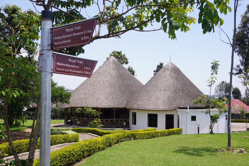 Two white huts with conical thatched roofs are surrounded by a well-kept lawn and small bushes. A post with two signs is on the left.