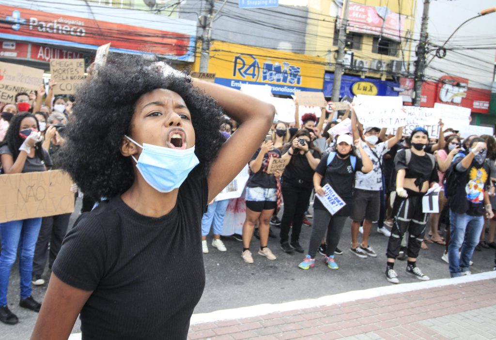 A young protester with curly dark hair and wearing a black shirt raises their fist in front of a group of people holding signs.