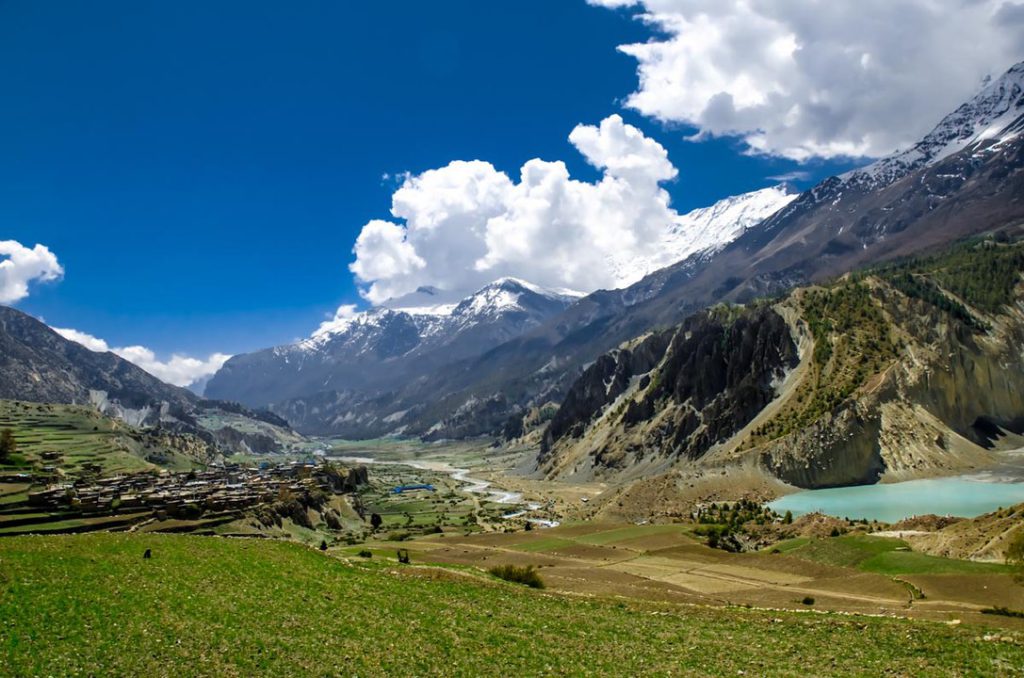 A wide shot shows a green valley with a blue pool of water surrounded by mountains below a bright blue sky with clouds.