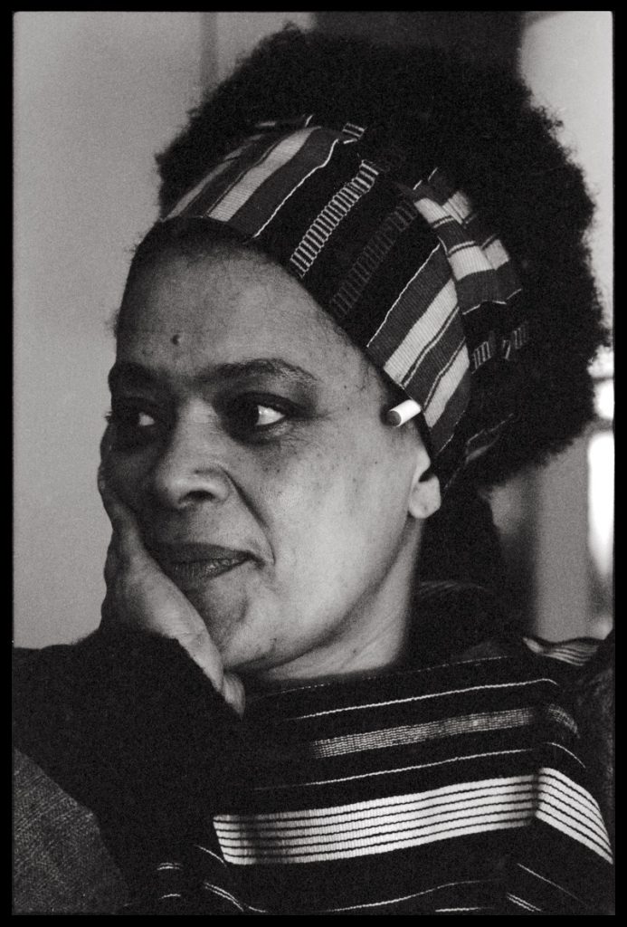 A black-and-white photo shows the portrait of a woman in a striped shirt and a striped head scarf and dark, curly hair looking sideways with her chin in the palm of her hand.