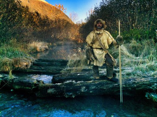 A man stands clothed in furs and tanned leather holding a staff beside a lush river landscape.