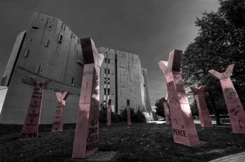 In a dramatic photograph, eight upright Y-shaped metal structures in red extend into the sky, each structure painted with an array of words and symbols reminiscent of Native American rock art.