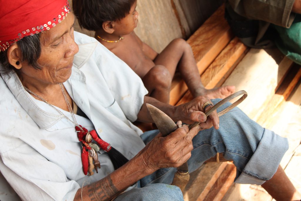 An elder wearing a red scarf on their head and a necklace with red and white pendants sits on a wooden bench next to a child.