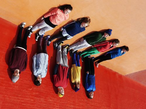 Two rows of people wearing colorful clothing lean against dark orange and light orange surfaces.