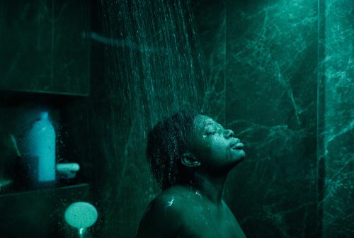 A person bathed in green light stands under falling water in front of marble walls with bottles on a shelf behind them.