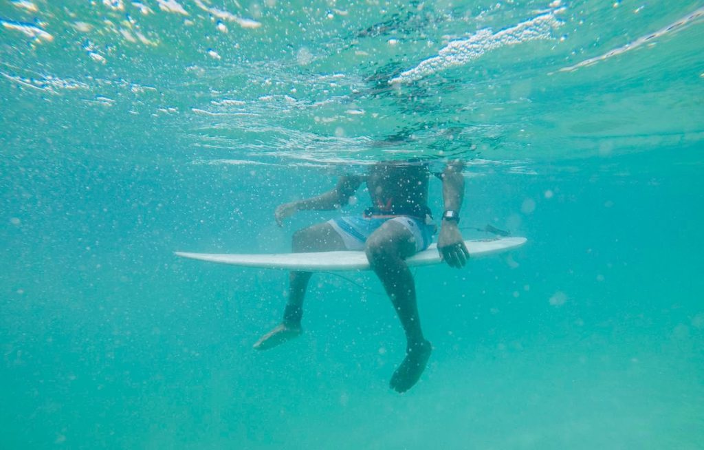 A person, seen from the chest downward from underwater, sits on a white surf board in turquoise water.