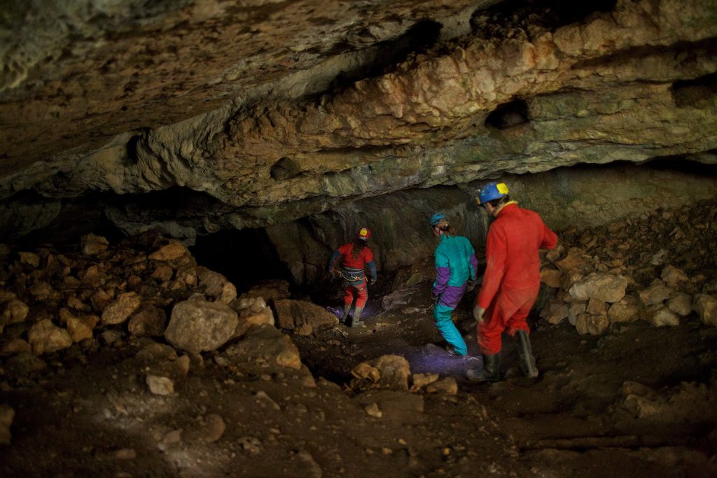 Three people in red and blue work suits, helmets, and boots walk in a cave.