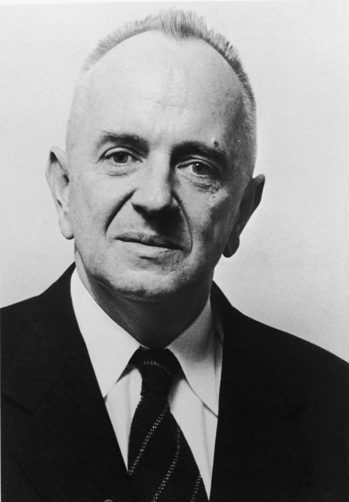 A black-and-white portrait shows a person in a suit and tie with short hair.