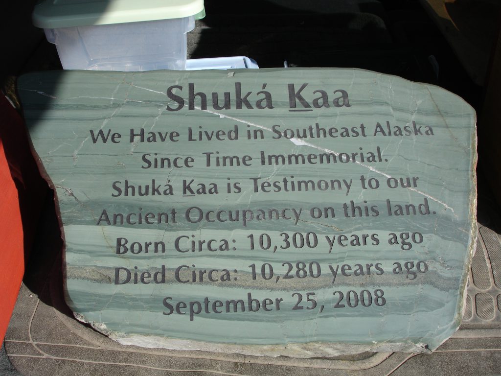 A slate-gray headstone dedicated to Shuká Káa sits in the back of a car.