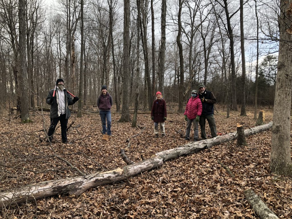 Five people dressed in winter clothes in the woods stand in a line, facing the camera and smiling.