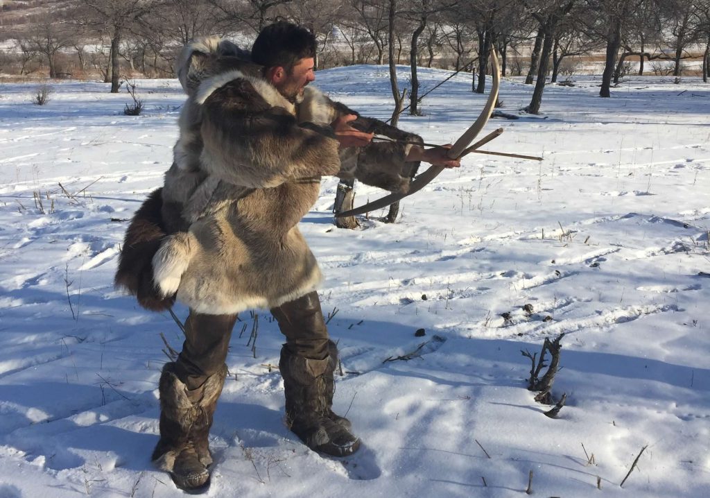 A man, dressed in hides and furs, pulls back an arrow on a wooden bow, taking aim.