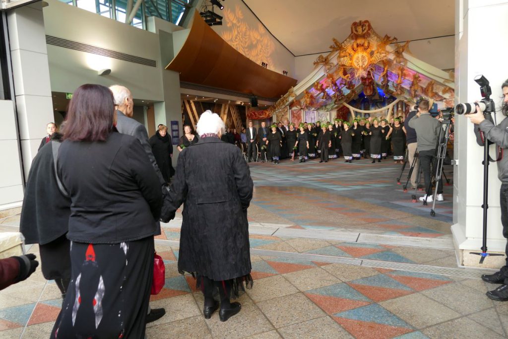 Five people enter a large museum room where a large groups of people face and greet them.