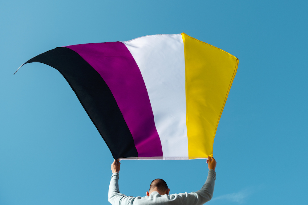 A person with a shaved head wearing a grey long-sleeved shirt holds a black, purple, white, and yellow striped flag over their head.