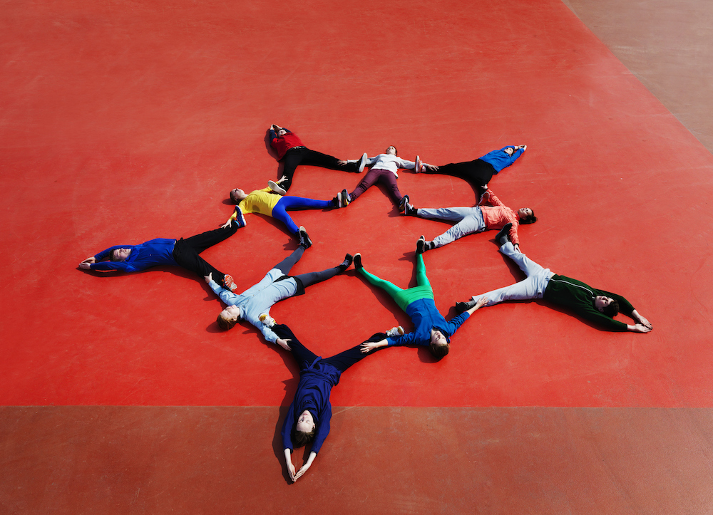 A group of people lie on an orange and brown floor, making a double star shape with their bodies.