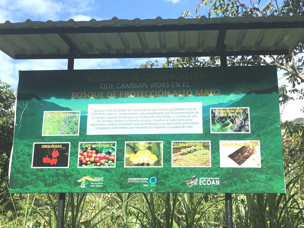 A large green sign featuring pictures of plants and text stands among tall grasses under an awning.