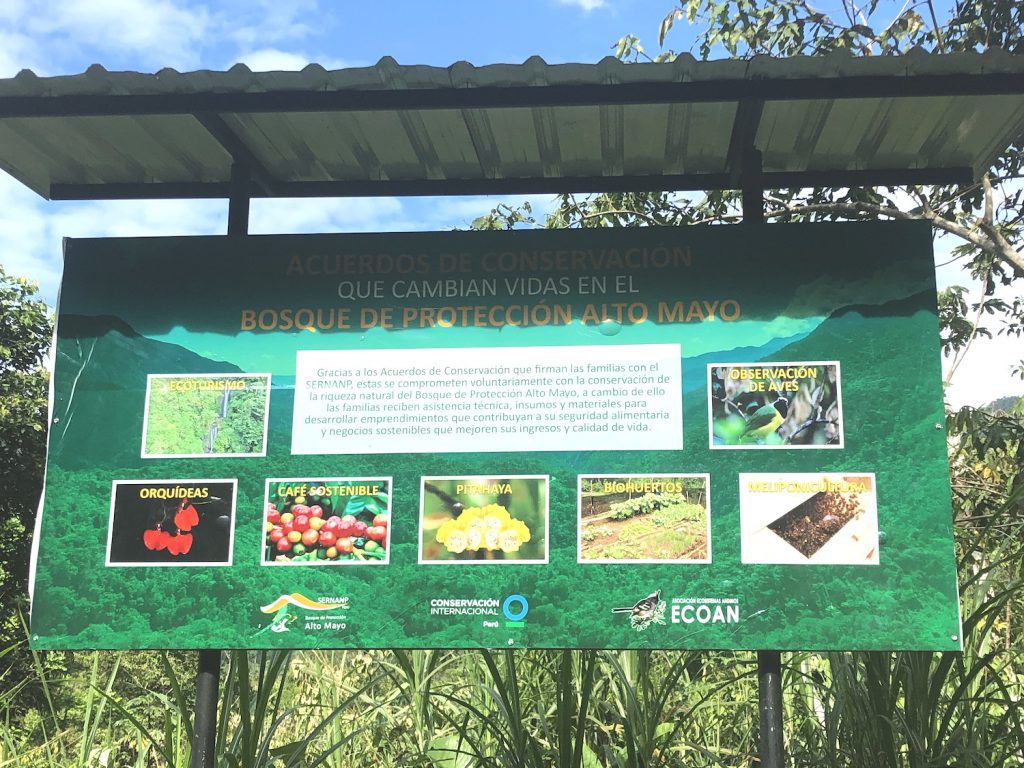 Un gran letrero verde con imágenes de plantas y texto se encuentra entre pastos altos debajo de un toldo.