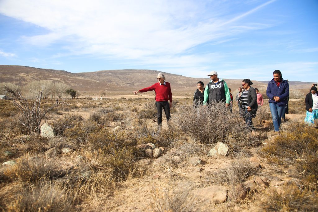 A person in a red shirt points out a spot to a group of people standing among dry grasses and rocks.