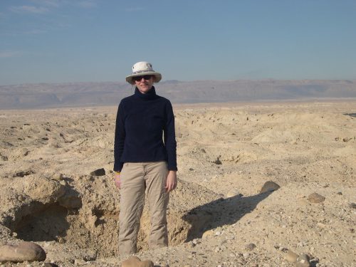 A person in a dark blue shirt, beige pants, and a wide-brimmed hat stands in a shallow sandy hole in the desert.