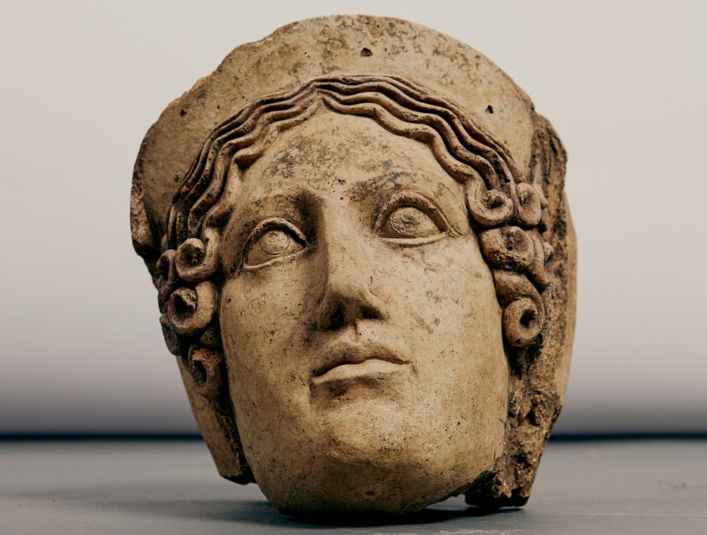 The head of a stone statue showing a stoic face and curly hair rests on a grey surface against a white background.