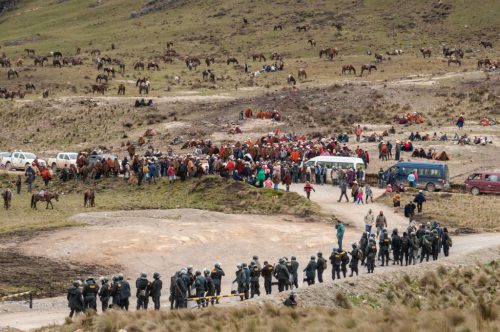 An aerial picture shows a hill covered with grazing animals, a large group of people in colorful clothing, and a line of armed police officers facing them.