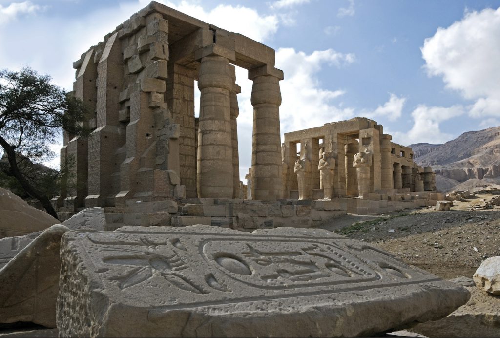 Two ancient stone buildings and columns stand on the sand under a blue sky with clouds. A broken slab of stone with hieroglyphs lies on the ground.