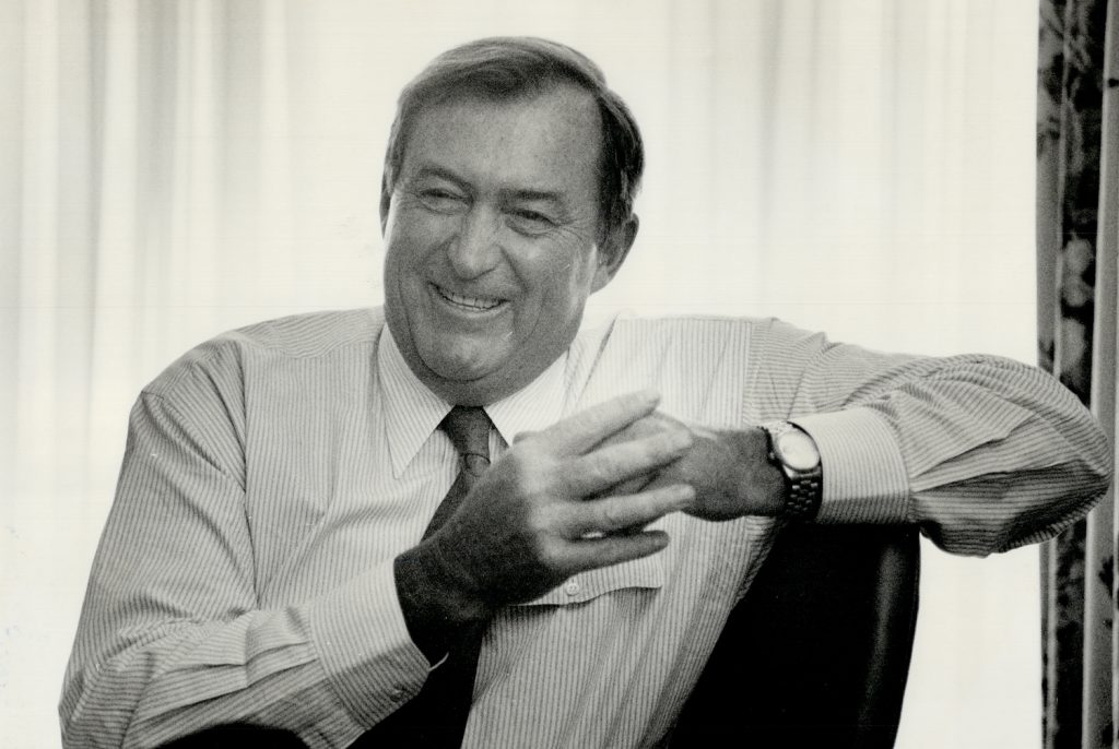 A person in a light-colored shirt and tie rests their arm on the back of a chair while pressing their hands together and smiling.