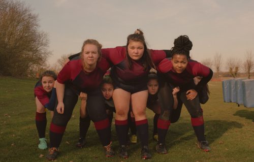 Members of a girls’ rugby team pose together with arms linked over each other’s shoulders, starting straight at the camera.