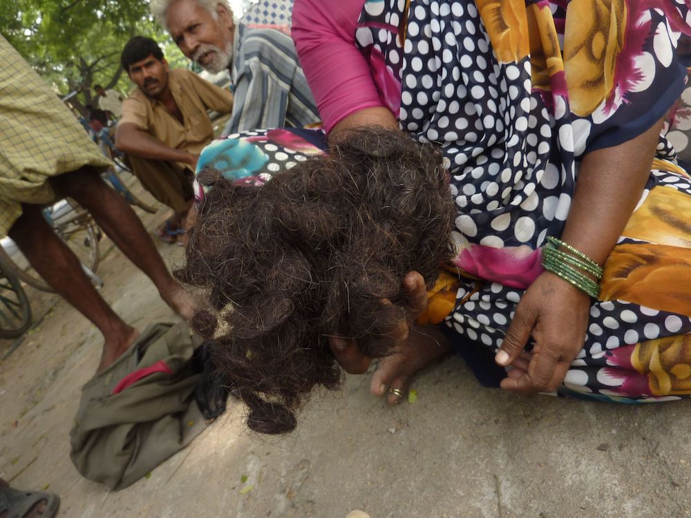 A person wearing a colorful dress and green bracelets sits on the ground and holds up a fistful of matted, dark hair.