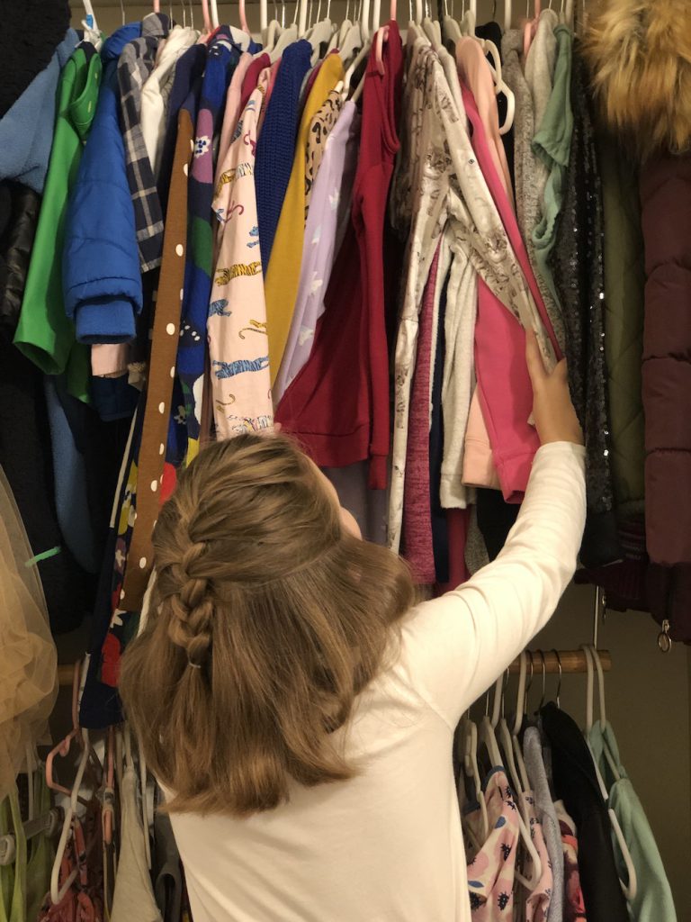 A girl with brown hair wearing a white shirt looks through clothing in a closet.