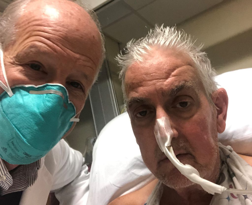 Two older men pose for a selfie, with one man wearing a surgical mask and the other with a tube extending from his nose.