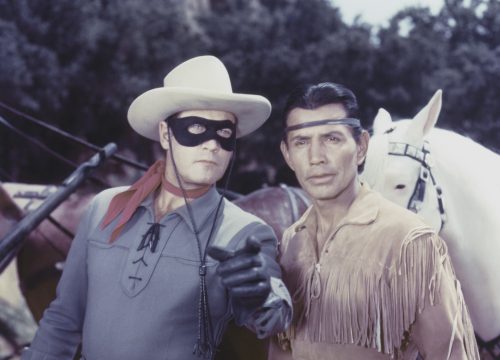 A person wearing a blue shirt, a red neck scarf, a black mask, and a white cowboy hat stands in front of horses and points to something in the distance. He is next to another person with wavy black hair wearing a brown shirt with fringe.
