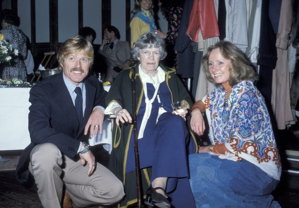 A person wearing a navy tie and jacket, and a person wearing a blue patterned shirt and blue jeans crouch down next to a seated person wearing a blue-and-white suit and holding a cane.