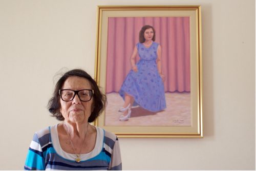 An older woman with short, brown hair and glasses wearing a blue, black, and white shirt stands in front of a portrait of a seated woman wearing a blue dress in front of pink curtains.