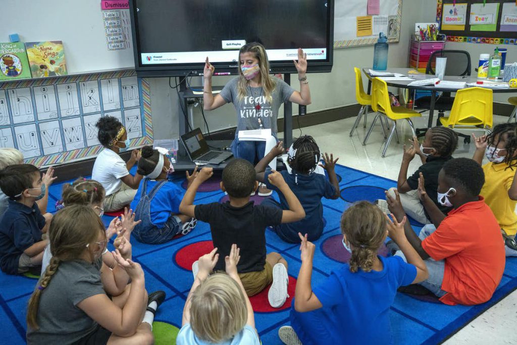 During the COVID-19 pandemic, an elementary teacher kneels on a blue mat in a classroom with a group of young children in front of them. They are all wearing masks and holding up their hands.