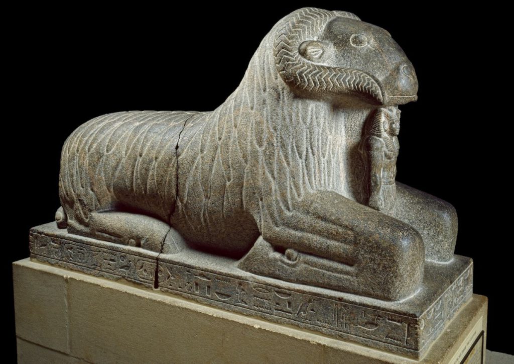 A gray stone statue displays a kneeling ram with curled horns and an Egyptian pharaoh standing between its front legs and under the ram’s chin against a black background.