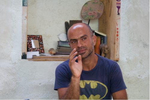 A person with a short, dark beard and sideburns wearing a navy-blue T-shirt with a yellow outline of the Batman symbol sits in front of a shelf with books and other items on it.